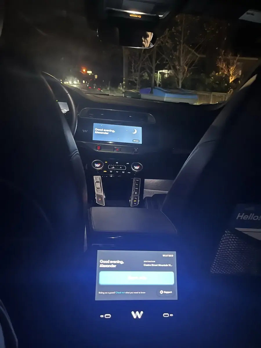 Inside a Waymo robotaxi: empty driver seat, passenger screen showing Good evening Alexander